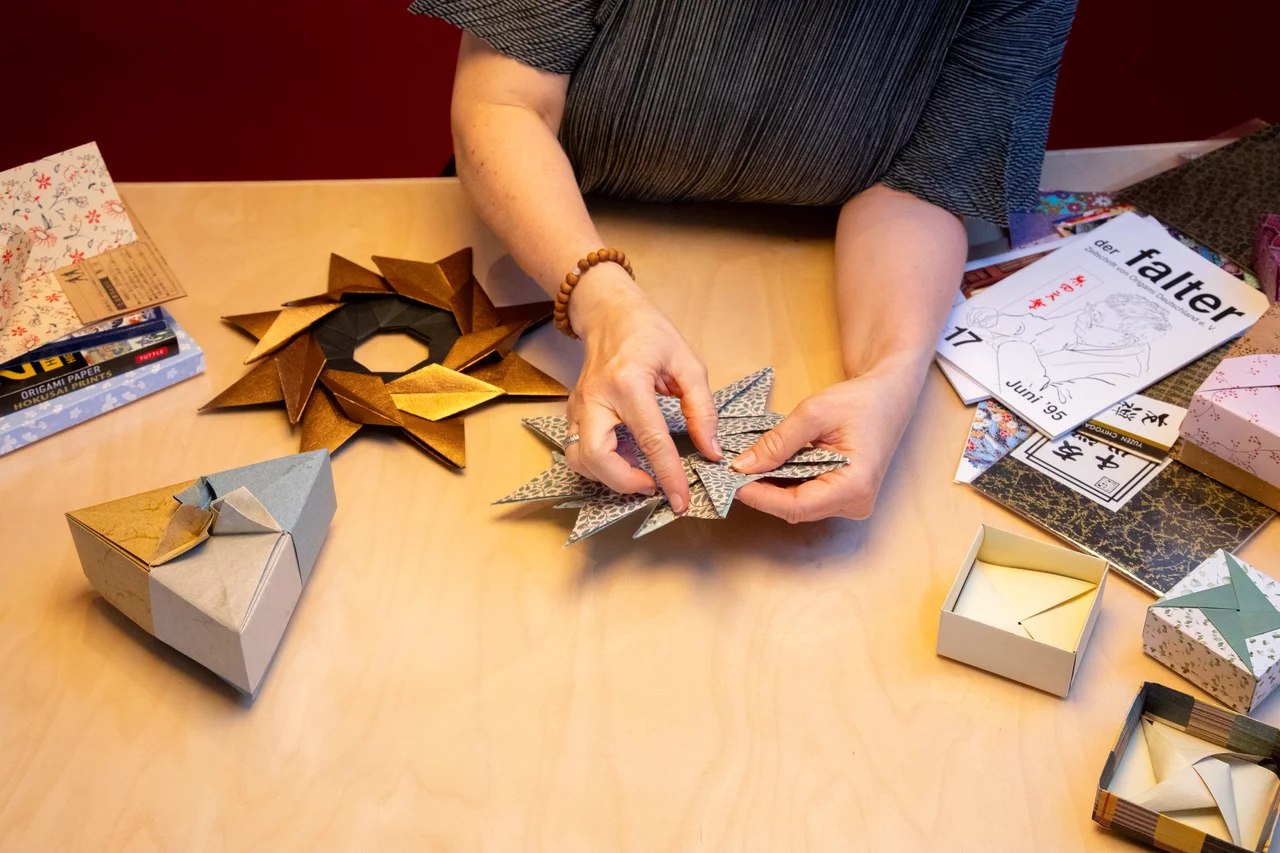 Ein Origamistern wird gerade zusammengesetzt. Daneben liegt ein Origamiblütenkranz und eine dreieckige Origamischachtel von Tomoko Fuse. Man sieht japanisches Origamipapier und den falter, die Vereinszeitschrift des Origamiverbands Deutschland. An der rechten Hand trägt die Künstlerin eine buddhistisches Mala, ein Armband aus Holzperlen.